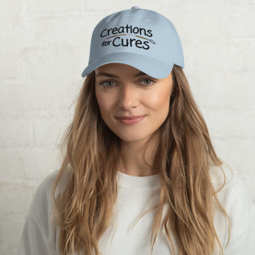 a person wearing a light blue "dad" hat featuring the Creations for Cures logo with the paintbrush in diverse skin tone colors