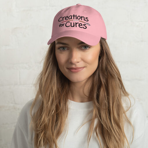 a person wearing a pink "dad" hat featuring the Creations for Cures logo with the paintbrush in diverse skin tone colors
