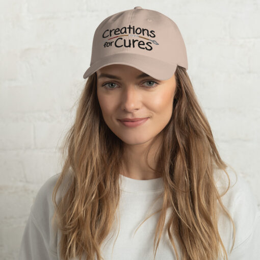 a person wearing a stone-color "dad" hat featuring the Creations for Cures logo with the paintbrush in diverse skin tone colors