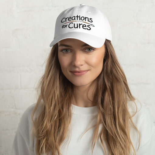 a person wearing a white "dad" hat featuring the Creations for Cures logo with the paintbrush in diverse skin tone colors