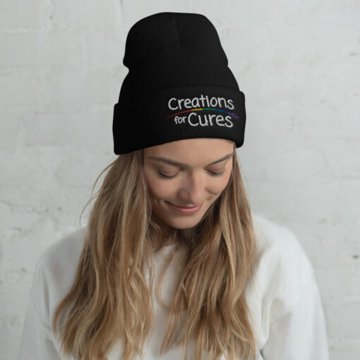 a person wearing a black beanie featuring the Creations for Cures logo with rainbow-striped paintbrush