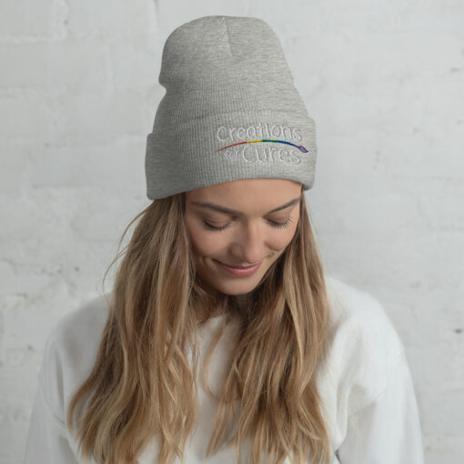 a person wearing a heather grey beanie featuring the Creations for Cures logo with rainbow-striped paintbrush