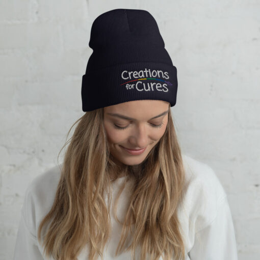 a person wearing a navy beanie featuring the Creations for Cures logo with rainbow-striped paintbrush