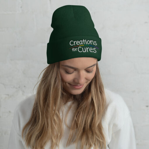 a person wearing a spruce green beanie featuring the Creations for Cures logo with rainbow-striped paintbrush