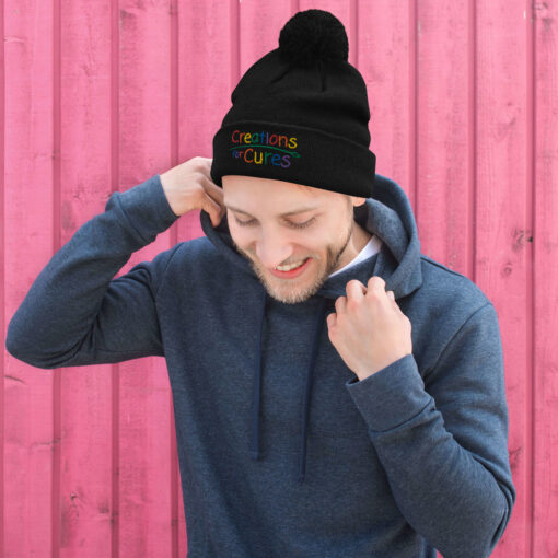 a person wearing a black pom pom knit cap featuring the Creations for Cures logo with rainbow lettering
