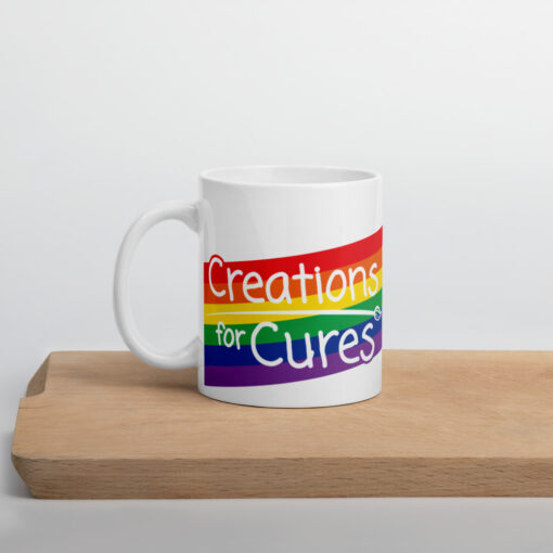 a white glossy mug featuring the Creations for Cures logo over a rectangular rainbow graphic