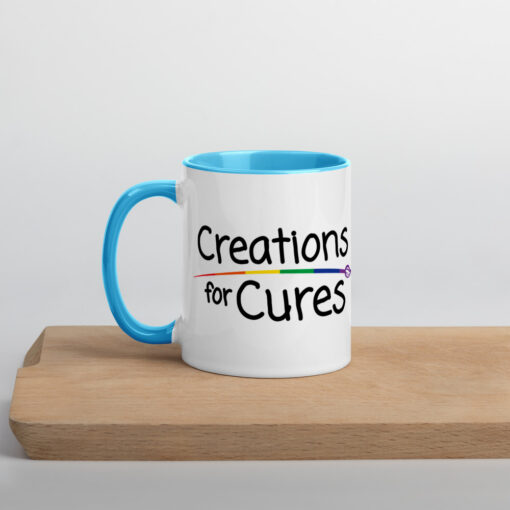 a white ceramic mug with blue handle and interior featuring the Creations for Cures logo