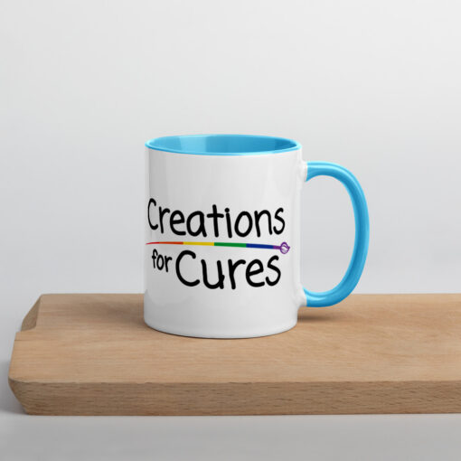 a white ceramic mug with blue handle and interior featuring the Creations for Cures logo