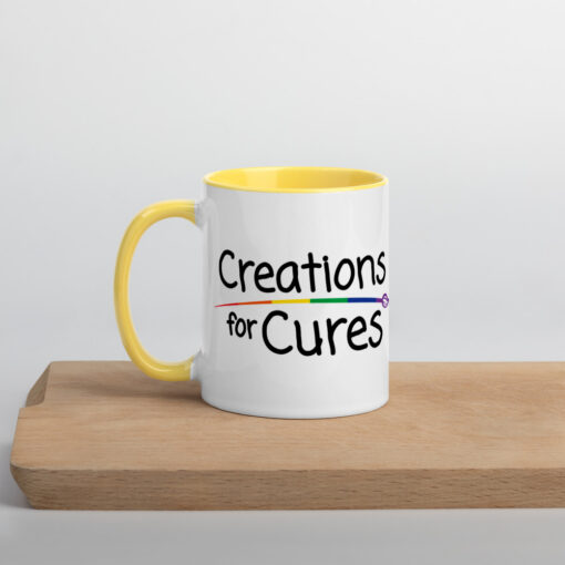 a white ceramic mug with yellow handle and interior featuring the Creations for Cures logo