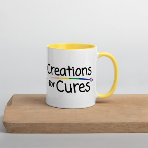 a white ceramic mug with yellow handle and interior featuring the Creations for Cures logo