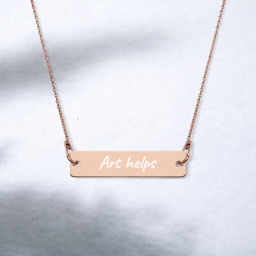 a rose gold bar necklace with the phrase "Art helps" engraved on it